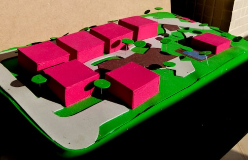 A colorful architectural model features several pink rectangular buildings on a green base, surrounded by circular green trees. Pavement paths are represented in gray, and there are various sections of different colors representing grass and other features.