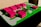 A colorful architectural model features several pink rectangular buildings on a green base, surrounded by circular green trees. Pavement paths are represented in gray, and there are various sections of different colors representing grass and other features.