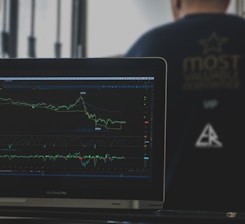 A laptop displays a detailed financial chart with lines showing data trends. In the background, a person is partially visible, wearing a dark shirt with the words 'most valuable' and some logos printed on it. The setting appears to be a modern workspace.