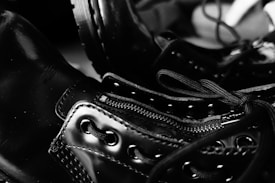 Close-up image of shiny black leather boots with detailed focus on the laces and zipper. The glossy surface of the boots reflects light, creating a contrast with the dark background. The stitching and eyelets are prominently visible, emphasizing the craftsmanship.