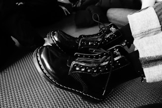 A pair of shiny black combat boots with detailed stitching and laces are worn by a person. The boots contrast with their striped pants, and they rest on a textured surface. Other people wearing dark clothing are visible in the background, partially blurred.