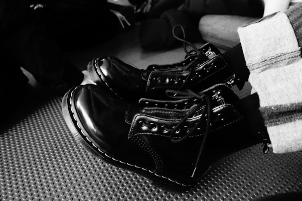 A pair of shiny black combat boots with detailed stitching and laces are worn by a person. The boots contrast with their striped pants, and they rest on a textured surface. Other people wearing dark clothing are visible in the background, partially blurred.