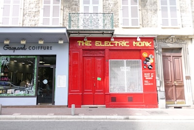A street view featuring two business facades and an adjacent door. On the left is a hair salon with a sign reading 'Gagpard Coiffure', showcasing a well-lit interior with hair products displayed. In the center, there is a brightly painted red storefront with the sign 'THE ELECTRIC MONK' in yellow lettering, decorated with vinyl records on the right side of its window, and information about buying and selling records. To the right, an imposing brown door is closed, embellished with a decorative lintel above.