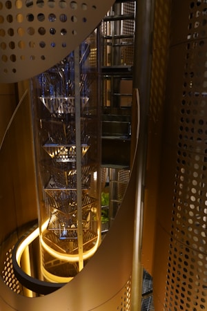 A futuristic elevator shaft with a sleek, metallic design encased in a cylindrical glass structure. The surrounding area features perforated metal panels with golden and bronze tones, creating an industrial yet elegant atmosphere. Ambient lighting highlights the geometric patterns and metallic surfaces, adding depth and dimension to the setting.