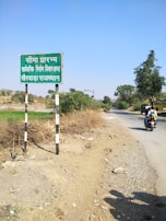 A rural road scene featuring a weathered sign with text in Hindi on the left, surrounded by dry shrubbery. A narrow paved road stretches into the distance on the right, bordered by greenery and scattered trees. A person on a motorcycle travels along the road, and the sky is clear with no clouds visible.