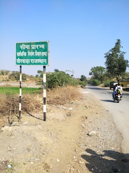 A rural road scene featuring a weathered sign with text in Hindi on the left, surrounded by dry shrubbery. A narrow paved road stretches into the distance on the right, bordered by greenery and scattered trees. A person on a motorcycle travels along the road, and the sky is clear with no clouds visible.