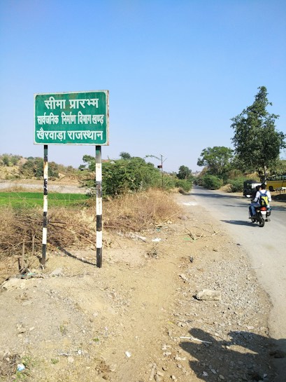A rural road scene featuring a weathered sign with text in Hindi on the left, surrounded by dry shrubbery. A narrow paved road stretches into the distance on the right, bordered by greenery and scattered trees. A person on a motorcycle travels along the road, and the sky is clear with no clouds visible.
