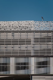 A modern building facade featuring a repetitive pattern composed of horizontal and vertical metallic slats. The upper part of the structure showcases a decorative panel with circular cutouts, adding a dynamic element against the backdrop of a clear blue sky. A light fixture is mounted atop the facade.