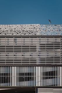 A modern building facade featuring a repetitive pattern composed of horizontal and vertical metallic slats. The upper part of the structure showcases a decorative panel with circular cutouts, adding a dynamic element against the backdrop of a clear blue sky. A light fixture is mounted atop the facade.