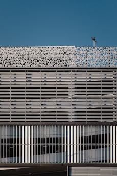 A modern building facade featuring a repetitive pattern composed of horizontal and vertical metallic slats. The upper part of the structure showcases a decorative panel with circular cutouts, adding a dynamic element against the backdrop of a clear blue sky. A light fixture is mounted atop the facade.