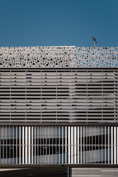 A modern building facade featuring a repetitive pattern composed of horizontal and vertical metallic slats. The upper part of the structure showcases a decorative panel with circular cutouts, adding a dynamic element against the backdrop of a clear blue sky. A light fixture is mounted atop the facade.