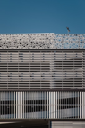 A modern building facade featuring a repetitive pattern composed of horizontal and vertical metallic slats. The upper part of the structure showcases a decorative panel with circular cutouts, adding a dynamic element against the backdrop of a clear blue sky. A light fixture is mounted atop the facade.
