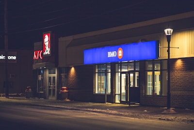 A street scene at night with dimly lit commercial buildings including a KFC and BMO Bank. The KFC sign is prominently visible with its red and white colors, while the BMO sign is illuminated in blue and orange. A street lamp casts a soft glow onto the sidewalk.