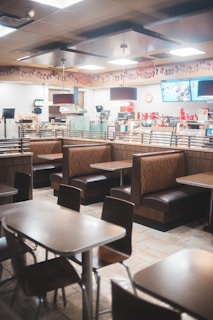 A fast food restaurant interior featuring empty tables and booths, with a modern design. The area is well-lit with overhead lighting and decorated with hanging decor. The counter area in the background includes digital menus and various appliances for food preparation.