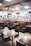 A fast food restaurant interior featuring empty tables and booths, with a modern design. The area is well-lit with overhead lighting and decorated with hanging decor. The counter area in the background includes digital menus and various appliances for food preparation.