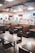 A fast food restaurant interior featuring empty tables and booths, with a modern design. The area is well-lit with overhead lighting and decorated with hanging decor. The counter area in the background includes digital menus and various appliances for food preparation.