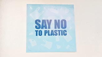 A light blue poster featuring bold, dark blue letters with the message 'Say No to Plastic'. The background has faint silhouettes of various plastic items like bottles and bags.