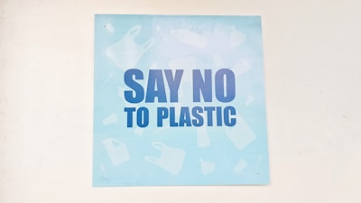 A light blue poster featuring bold, dark blue letters with the message 'Say No to Plastic'. The background has faint silhouettes of various plastic items like bottles and bags.