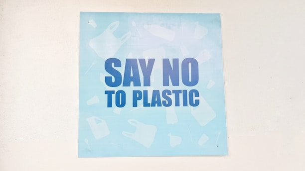 A light blue poster featuring bold, dark blue letters with the message 'Say No to Plastic'. The background has faint silhouettes of various plastic items like bottles and bags.