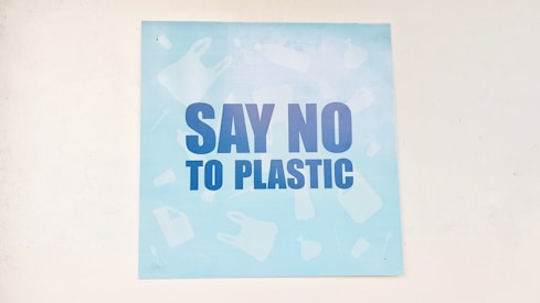 A light blue poster featuring bold, dark blue letters with the message 'Say No to Plastic'. The background has faint silhouettes of various plastic items like bottles and bags.