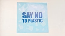 A light blue poster featuring bold, dark blue letters with the message 'Say No to Plastic'. The background has faint silhouettes of various plastic items like bottles and bags.