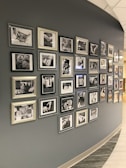 A curved wall displaying numerous framed black-and-white photographs and text. The photographs appear to be close-ups of hands and possibly people in different settings, with some images accompanied by inspirational or informational text. The frames vary in style and size, adding an eclectic yet organized feel to the display.