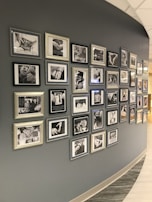 A curved wall displaying numerous framed black-and-white photographs and text. The photographs appear to be close-ups of hands and possibly people in different settings, with some images accompanied by inspirational or informational text. The frames vary in style and size, adding an eclectic yet organized feel to the display.
