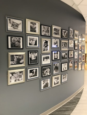 A curved wall displaying numerous framed black-and-white photographs and text. The photographs appear to be close-ups of hands and possibly people in different settings, with some images accompanied by inspirational or informational text. The frames vary in style and size, adding an eclectic yet organized feel to the display.