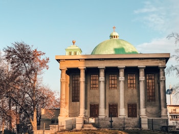 A building with classical architecture, featuring tall columns and a domed roof with a cross on top. The walls are accented with wooden window panels. Autumn trees with orange and brown leaves surround the structure, under a clear blue sky.