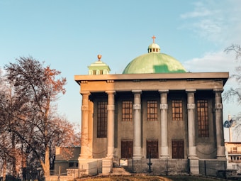 A building with classical architecture, featuring tall columns and a domed roof with a cross on top. The walls are accented with wooden window panels. Autumn trees with orange and brown leaves surround the structure, under a clear blue sky.