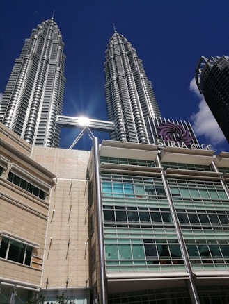 A photograph features the iconic Petronas Twin Towers against a clear blue sky, with the sun reflecting off the structure. In the foreground, the Suria KLCC shopping mall is visible, displaying its modern architectural design with glass windows and a beige facade.