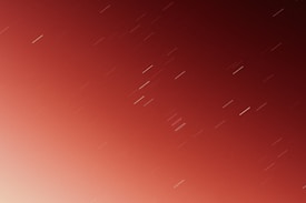 A long-exposure photograph capturing the night sky with visible star trails. The colors transition from a deep red at the top to a lighter hue at the bottom, creating a gradient effect. The star trails are diagonal, indicating the passage of time during the exposure.