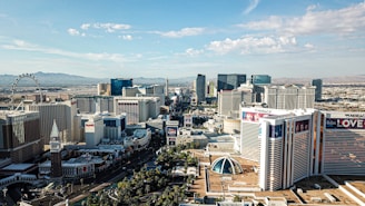 Aerial view of a cityscape featuring prominent buildings, hotels, resorts, and casinos. In the background, there is a Ferris wheel, and distant mountains are visible on the horizon. Some of the buildings have large billboards advertising various shows and attractions.