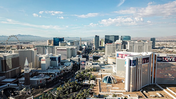 Aerial view of a cityscape featuring prominent buildings, hotels, resorts, and casinos. In the background, there is a Ferris wheel, and distant mountains are visible on the horizon. Some of the buildings have large billboards advertising various shows and attractions.