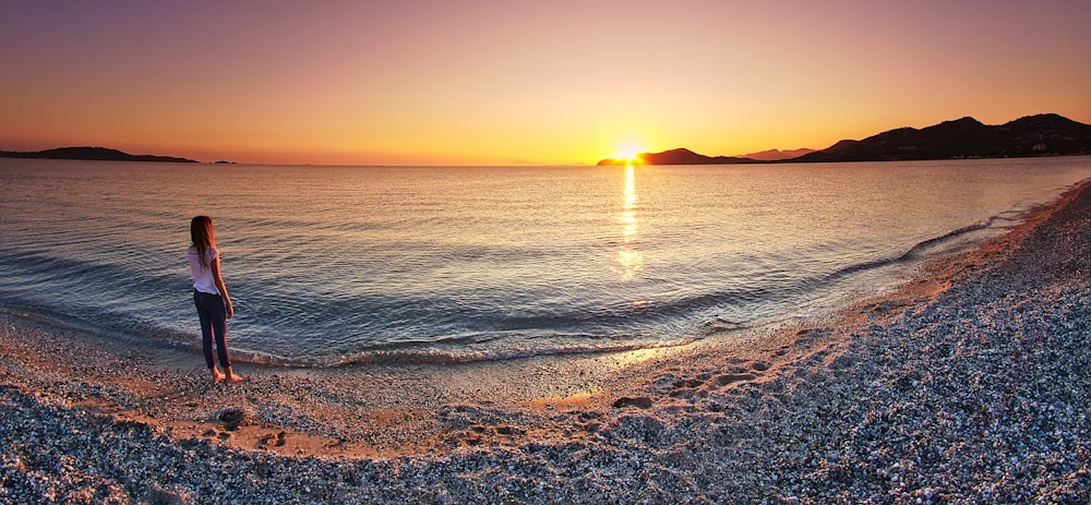 woman wearing white top standing and facing on body of water under orange sky