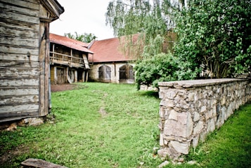 A rustic, countryside scene with two wooden and stone buildings featuring red-tiled roofs. A grassy area is in the foreground, bordered by a stone wall and lush green bushes. The weathered wood and stone construction gives a quaint, historical feel.