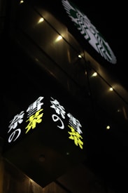 A nighttime scene featuring a building adorned with illuminated signage. The signage includes a Japanese text in a bright yellow and white color against a dark background. Above, there's a round, lighted logo with a design that is partly visible.