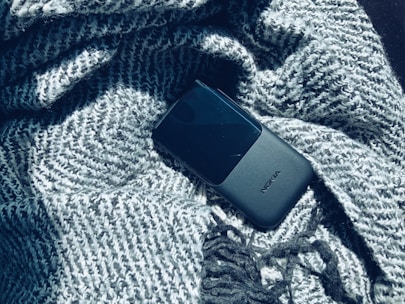A black mobile phone with the brand name visible lies on a textured grey knitted fabric, partially wrapped and casting soft shadows.