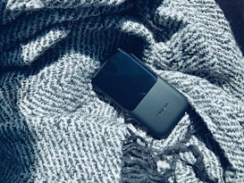 A black mobile phone with the brand name visible lies on a textured grey knitted fabric, partially wrapped and casting soft shadows.