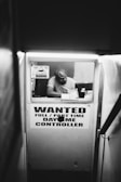 A narrow passageway leads to a small booth with a person inside wearing a hat, looking at some papers. On the booth window, there is a large sign that reads 'WANTED FULL / PART TIME DAY/NIGHT CONTROLLER' and a smaller 'WARNING' sign.