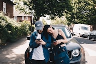 A young couple is standing next to a parked car on a sunny street. Both are looking at their phones, with the woman leaning her head on the man's shoulder. The street is lined with trees and there is a white van and another car parked along the curb.