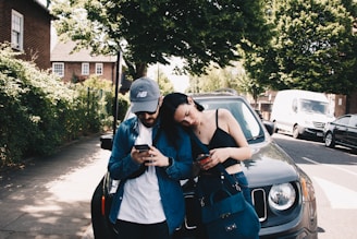 A young couple is standing next to a parked car on a sunny street. Both are looking at their phones, with the woman leaning her head on the man's shoulder. The street is lined with trees and there is a white van and another car parked along the curb.