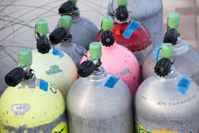 A collection of scuba diving tanks is arranged closely together. The tanks have various colors including yellow, red, blue, gray, and pink. They are fitted with black valves and green caps, and are marked with blue labels or tags.