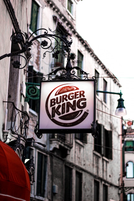 A decorative sign displaying the Burger King logo hangs from an ornate wrought iron bracket against the backdrop of an old stone building with green shuttered windows. The scene suggests an urban environment with a mix of historical and modern elements.