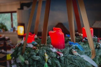 A decorative wreath with bright red candles arranged among lush green foliage, accented with various small ornaments and feathers. Soft lighting in the background creates a cozy, warm atmosphere.