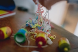 Colorful party popper confetti spread across a wooden surface with a person's hand holding some strands. Several poppers are scattered around, partially opened with visible bright colors.