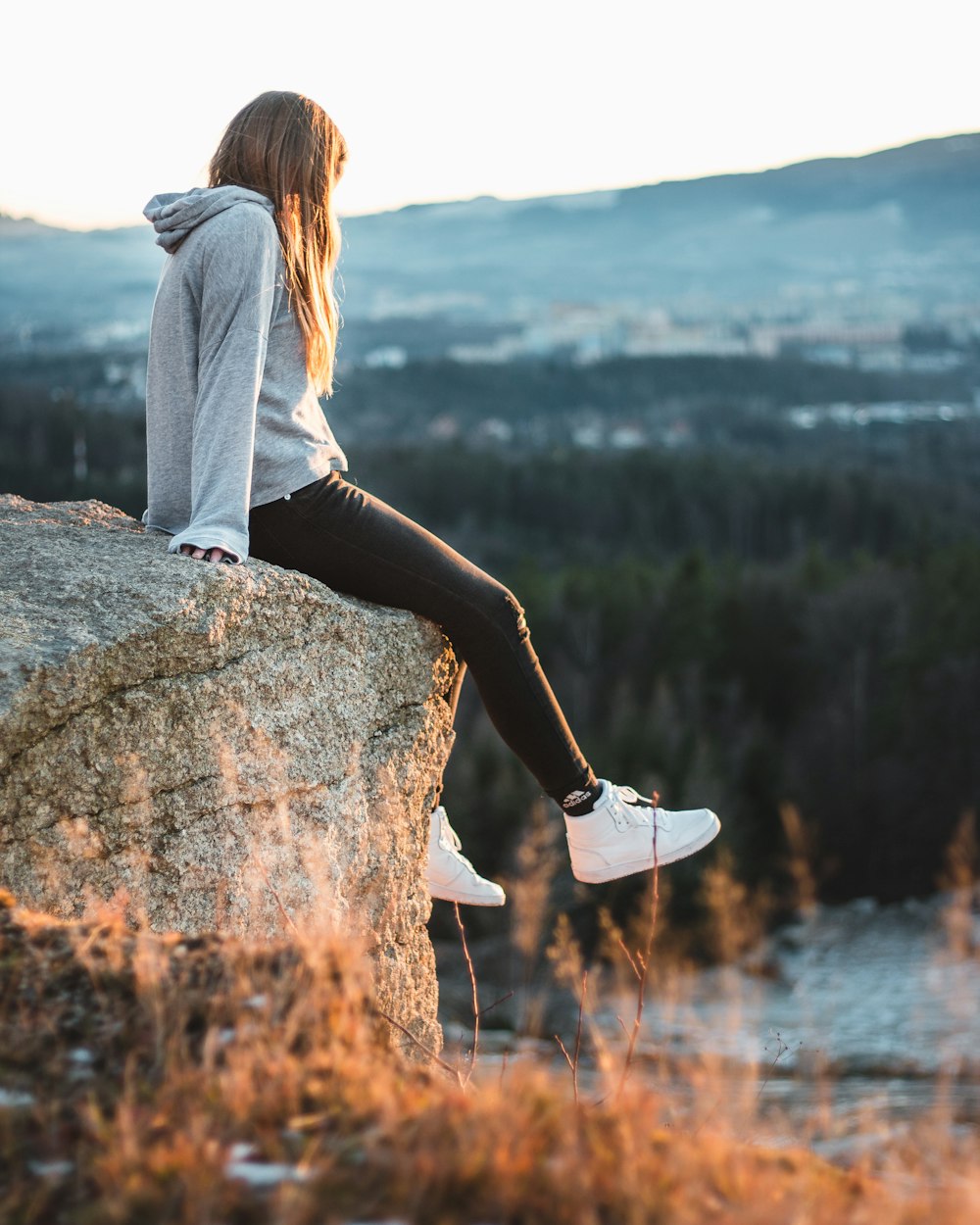 woman in gray hoodie and black pants sitting on rock cliff