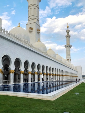 A grand mosque with large white domes and intricate gold decorations. The building features extensive arches and pillars. A reflective pool sits in the foreground, bordered by a well-maintained lawn. The sky is partly cloudy, adding to the serene atmosphere.