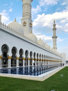 A grand mosque with large white domes and intricate gold decorations. The building features extensive arches and pillars. A reflective pool sits in the foreground, bordered by a well-maintained lawn. The sky is partly cloudy, adding to the serene atmosphere.