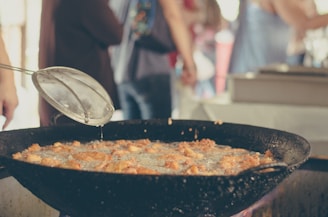 A close-up view of a large pan filled with oil where pieces of food are being deep-fried. A skimmer is held above the pan, allowing excess oil to drip back into it. In the background, blurred figures of people suggest a busy environment, possibly a market or festival.